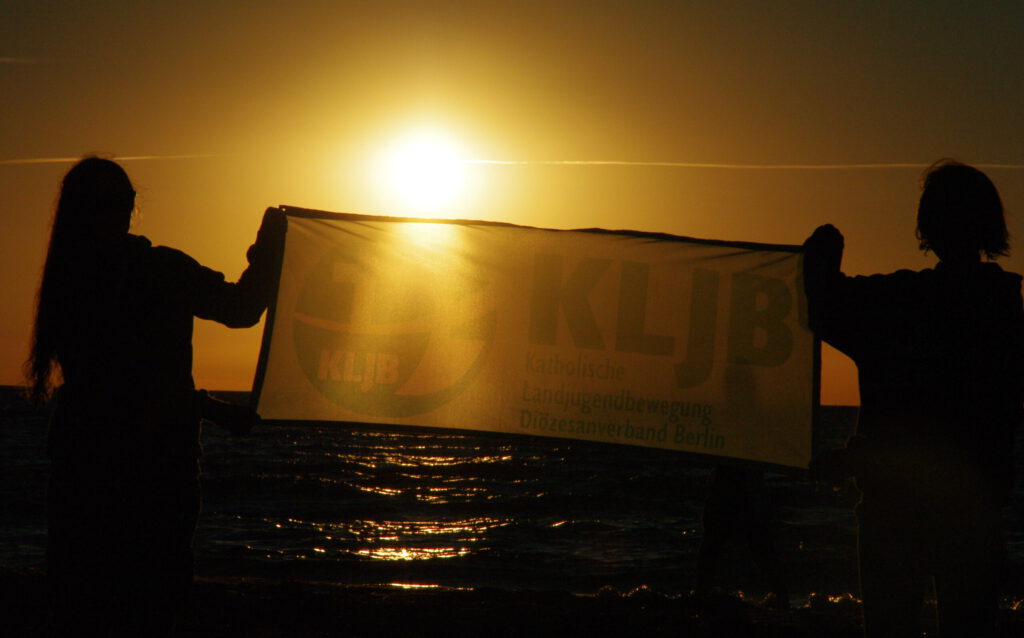 zwei Menschen halten ein Banner mit KLJB Logo hoch. Im Hintergrund sieht man den Sonnenuntergang überm Meer.